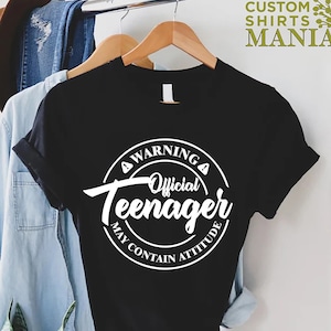 May include: Black t-shirt with a white circle graphic that says "Warning Official Teenager May Contain Attitude".