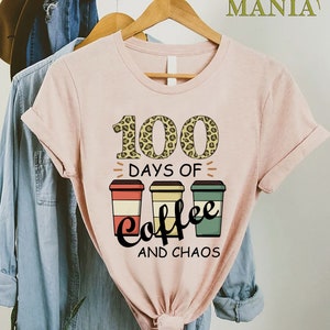 Pu&ograve; includere: Maglietta rosa con un design grafico di tre tazze di caff&egrave; e il testo "100 giorni di caff&egrave; e caos". Il numero 100 &egrave; decorato con uno stampa leopardato.