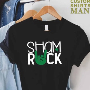 Peut inclure: T-shirt noir avec du texte blanc qui dit "Shamrock" et "Rock" avec une main verte faisant le signe rock and roll.