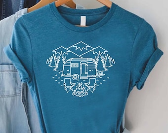 May include: A teal blue t-shirt with a white outline design of a camper van parked in a forest setting with mountains in the background. The design includes trees, a campfire, and two chairs.