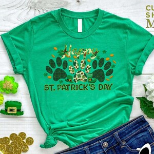 May include: Green t-shirt with a paw print design and the text "Happy St. Patrick's Day". The shirt is on a white wooden surface with a pair of denim shorts, gold coins, and a green shamrock.