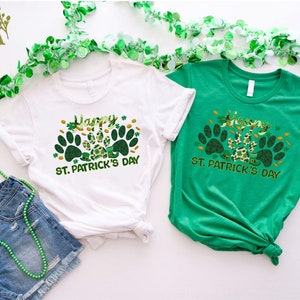 May include: Two t-shirts with a green shamrock and paw print design. The white t-shirt says "Happy St. Patrick's Day" and the green t-shirt says "Happy St. Patrick's Day".