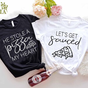 Puede incluir: Dos camisetas blancas y negras con un gr&aacute;fico de pizza y texto. La camiseta negra dice "Se rob&oacute; una pizza de mi coraz&oacute;n" y la camiseta blanca dice "Vamos a emborracharnos".