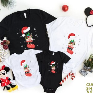 May include: Set of four Christmas-themed t-shirts featuring a stocking filled with presents and a Santa hat. The shirts are black and white with the words "Daddy", "Mama", "Name", and "Name" on each shirt.