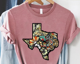 May include: A pink t-shirt with a colorful, doodle-style outline of the state of Texas. The design features flowers, birds, and other whimsical elements.