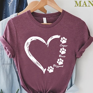 May include: A maroon t-shirt with a white heart and paw prints. The text "Cooper, Roxie, Blizzard" is written below the paw prints.