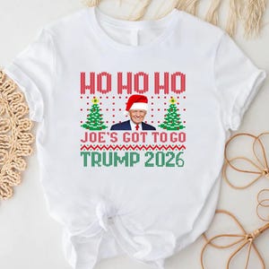 Op de afbeelding: Een wit T-shirt met korte mouwen en een grafische print in rood, groen en blauw. Het ontwerp bevat de tekst HO HO HO, JOE'S GOT TO GO en TRUMP 2026, samen met twee groene kerstbomen en een portret van Donald Trump met een kerstmuts op.