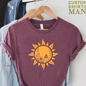 Puede incluir: Una camiseta de color granate con un gr&aacute;fico de sol amarillo. El sol tiene un dise&ntilde;o floral con mariposas. La camiseta est&aacute; en una percha.