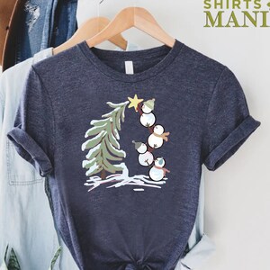 May include: A dark blue t-shirt with a graphic of snowmen climbing a Christmas tree. The snowmen are wearing hats and scarves. The tree is green with white snow on the branches.