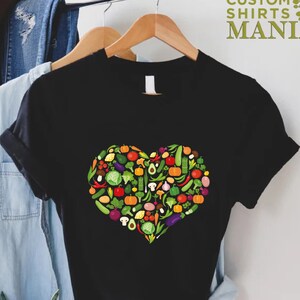 May include: Black t-shirt featuring a heart-shaped design filled with colorful vegetables. The vegetables include tomatoes, peppers, avocados, and more. The text "CUSTOM SHIRTS MANIA" is printed in green on the upper right.