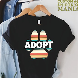 May include: Black t-shirt with a colorful retro-style paw print and the word "ADOPT" in white text.