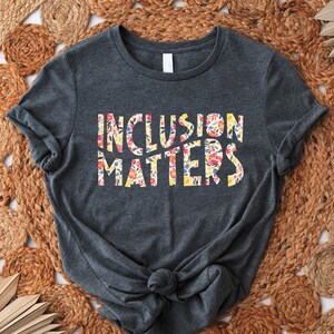 Puede incluir: Una camiseta gris oscuro con el texto "Inclusion Matters" impreso en letras blancas con un patr&oacute;n floral. La camiseta est&aacute; colocada sobre una estera de paja tejida.