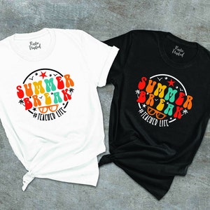 May include: Two t-shirts with a retro-style graphic design. The white t-shirt has a red, orange, yellow, green, and blue graphic that says "Summer Break" with sunglasses and the hashtag "#TeacherLife". The black t-shirt has the same graphic in the same colors.