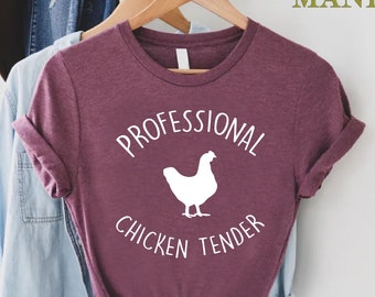 Könnte beinhalten: Burgunderfarbenes T-Shirt mit hochgekrempelten Ärmeln. Der weiße Text "PROFESSIONAL CHICKEN TENDER" und eine weiße Hühner-Grafik sind aufgedruckt. Das T-Shirt hängt an einem Holzbügel. Teil einer Bekleidungskollektion.