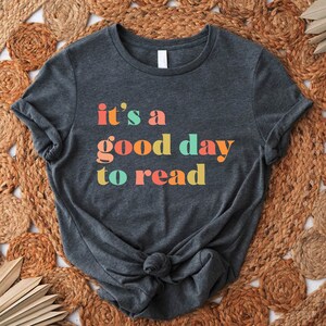 May include: A dark gray t-shirt with the text "it's a good day to read" in colorful letters. The letters are in shades of orange, yellow, green, and blue.