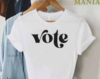 May include: White t-shirt with the word "vote" printed in black, stylized lettering. The shirt is hanging on a wooden hanger.