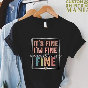 May include: Black t-shirt with a white box around the text "It's fine I'm fine everything's fine" in rainbow colors.