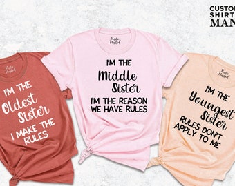 Puede incluir: Tres camisetas con diferentes lemas sobre ser hermana. La primera camiseta es de color óxido y dice "Soy la hermana mayor, yo pongo las reglas". La segunda camiseta es rosa y dice "Soy la hermana del medio, soy la razón por la que hay reglas". La tercera camiseta es de color melocotón y dice "Soy la hermana menor, las reglas no se aplican a mí".