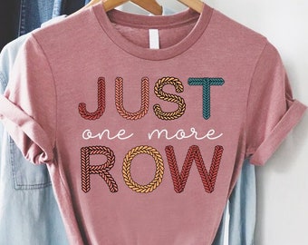 May include: A dusty rose t-shirt with the text "JUST one more ROW" in a knitted font. The letters are in shades of orange, yellow, and teal. The shirt is hanging on a wooden hanger with a blue denim shirt and a hat in the background.