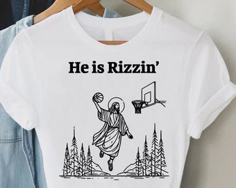 May include: White t-shirt with a black and white illustration of a person playing basketball with the text "He is Rizzin'" printed on it.