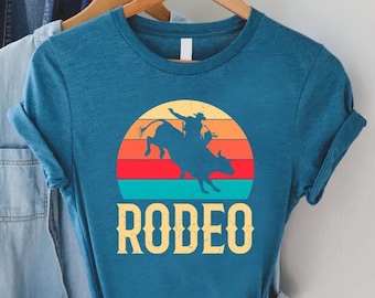 Könnte beinhalten: Ein blaues T-Shirt mit einem Vintage-Sonnenuntergang-Motiv, das einen Cowboy auf einem Stier zeigt. Der Text "RODEO" ist unter dem Motiv gedruckt.