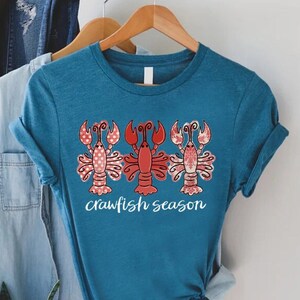 May include: Teal t-shirt featuring three cartoon crawfish in red and pink patterns. The middle crawfish is solid red. The words "crawfish season" are written in white script below the crawfish. The shirt is hanging on a wooden hanger.