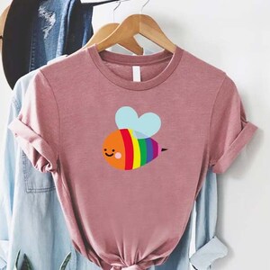 May include: A pink t-shirt with a rainbow bee graphic. The bee has a smiling face and a light blue heart-shaped wing.