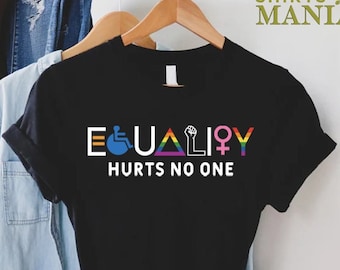 May include: Black t-shirt with the word "EQUALITY" in white, featuring symbols for disability, LGBTQ+, and gender equality. The text "HURTS NO ONE" is below. The shirt is hanging on a wooden hanger.