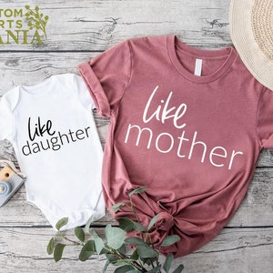 以下が含まれることがあります： 白とピンクの2枚のシャツのセットで、それぞれ「like daughter」と「like mother」の文字がプリントされています。シャツは木製の表面に置かれています。