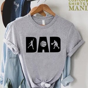 Op de afbeelding: Grijs T-shirt met het woord "DAD" in zwart met een basketbal en twee basketballers in de letters.