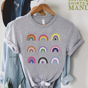 May include: A gray t-shirt with a design of nine different colored rainbows. The rainbows are arranged in three rows of three. The text "CUSTOM SHIRTS MANIA" is in the top right corner of the image.