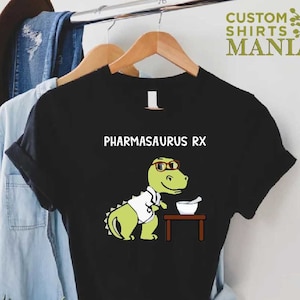 May include: Black t-shirt featuring a green dinosaur wearing glasses and a white lab coat. The dinosaur is holding a mortar and pestle. The text "PHARMASAURUS RX" is printed above the dinosaur.
