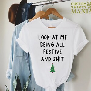 May include: White t-shirt with black text that reads "Look at me being all festive and shit" with a green Christmas tree graphic below the text.