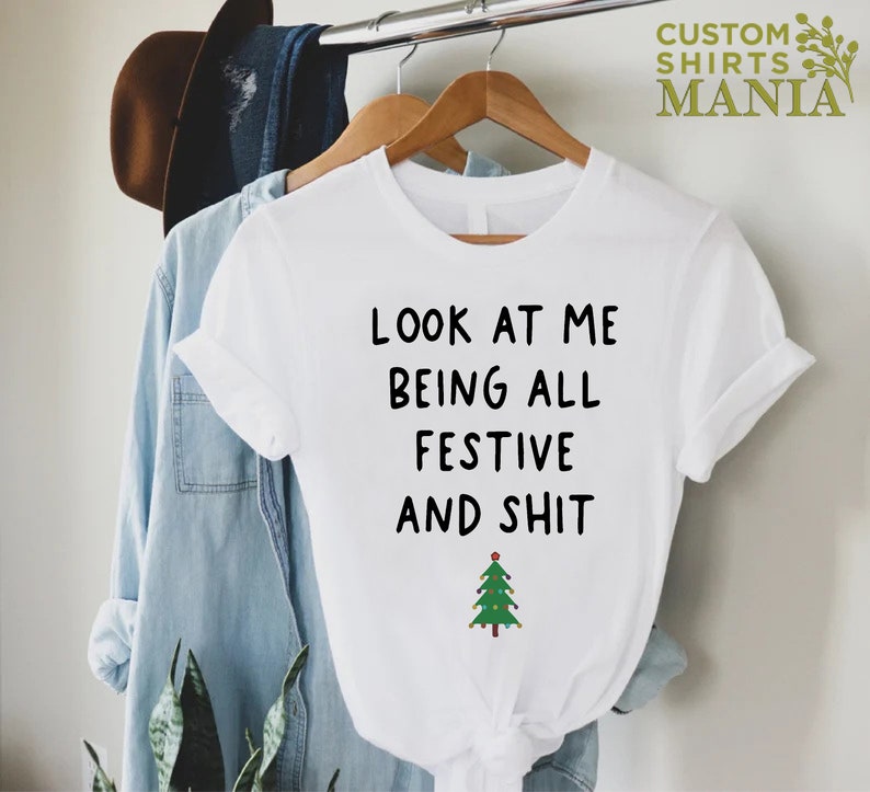 May include: White t-shirt with black text that reads "Look at me being all festive and shit" with a green Christmas tree graphic below the text.