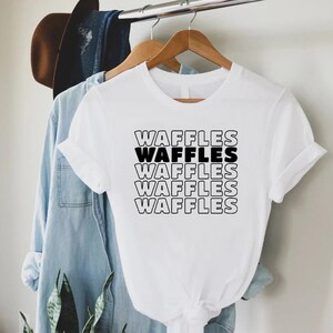 Może przedstawiać: Biała koszulka z napisem "WAFFLES" powt&oacute;rzonym pięć razy w stosie, pogrubioną czarną czcionką z białym obramowaniem.