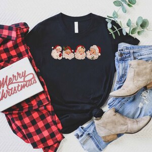 May include: A black t-shirt with a graphic of four vintage Santa Claus faces. The shirt is laid out on a white surface with a red and black plaid shirt, a pair of blue jeans, and a pair of brown ankle boots.