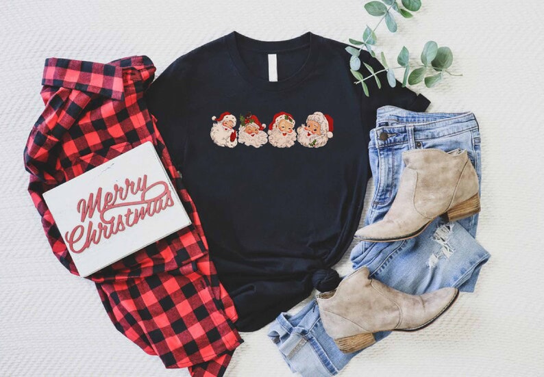 May include: A black t-shirt with a graphic of four vintage Santa Claus faces. The shirt is laid out on a white surface with a red and black plaid shirt, a pair of blue jeans, and a pair of brown ankle boots.