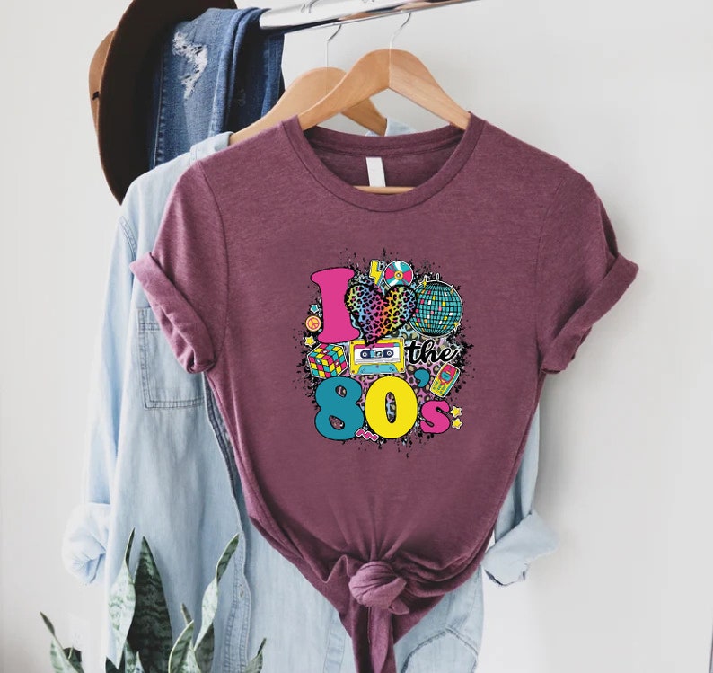 May include: A maroon t-shirt with a graphic print of a heart with a leopard print, a disco ball, a cassette tape, a Rubik's cube, and the text "I love the 80s".