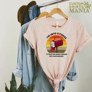 May include: A pink t-shirt with a retro-style graphic of a red mailbox with the text "I'm into fitness. Fit'ness this whole package into your mailbox."