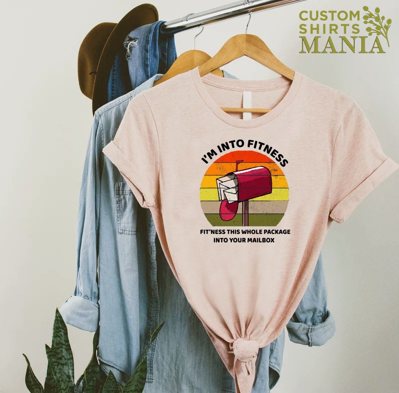 May include: A pink t-shirt with a retro-style graphic of a red mailbox with the text "I'm into fitness. Fit'ness this whole package into your mailbox."
