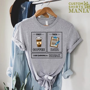 May include: Gray t-shirt with a graphic of two boxes. The first box says "First - Coffee" and has a cartoon image of a cup of coffee. The second box says "Then - Data" and has a cartoon image of a clipboard and pen. The text below the boxes says "I am earning a: Break".