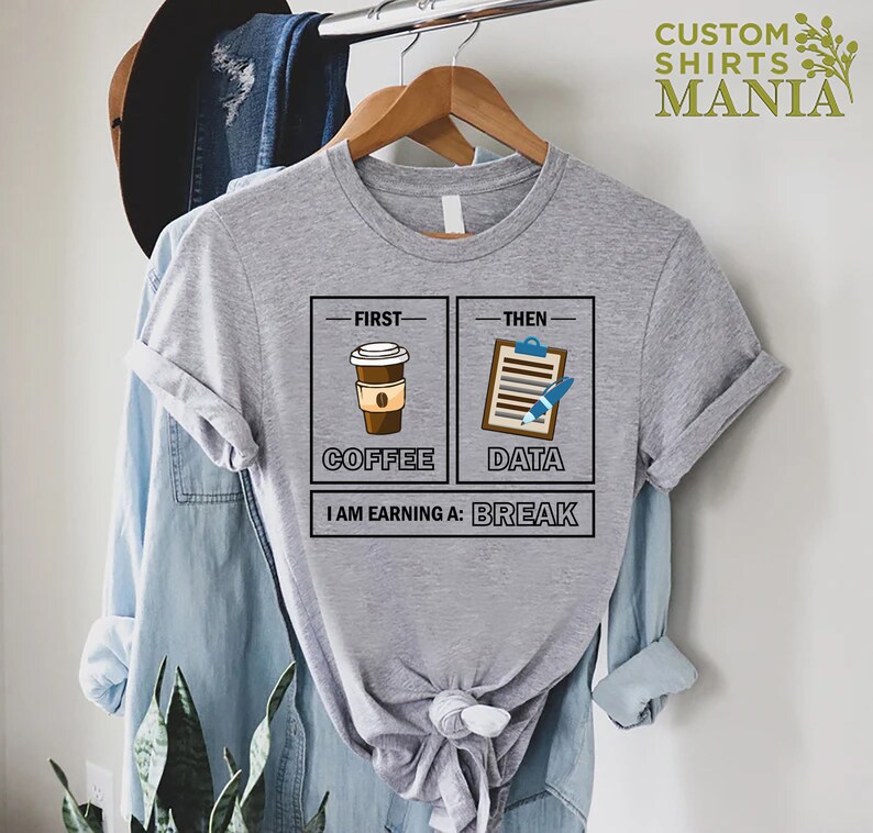 May include: Gray t-shirt with a graphic of two boxes. The first box says "First - Coffee" and has a cartoon image of a cup of coffee. The second box says "Then - Data" and has a cartoon image of a clipboard and pen. The text below the boxes says "I am earning a: Break".