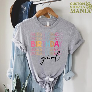 May include: A grey t-shirt with the text "Birthday girl" in rainbow colours. The text is stacked and repeated multiple times.