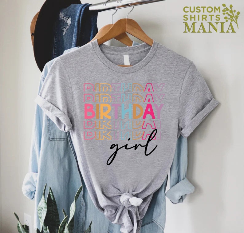 May include: A grey t-shirt with the text "Birthday girl" in rainbow colours. The text is stacked and repeated multiple times.