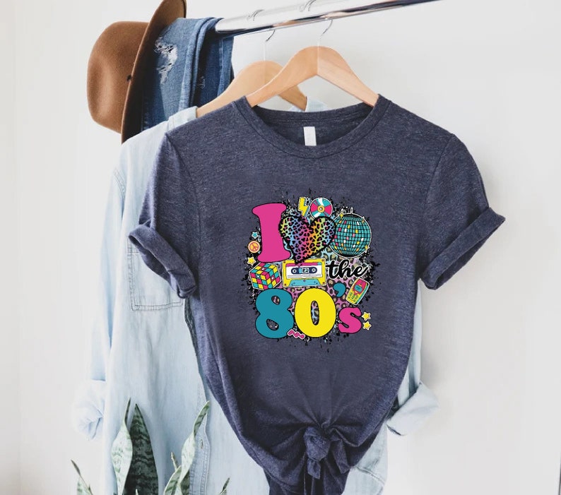 May include: A dark blue t-shirt with a colorful graphic design featuring a heart with a leopard print, a cassette tape, a disco ball, and the text "I love the 80's".
