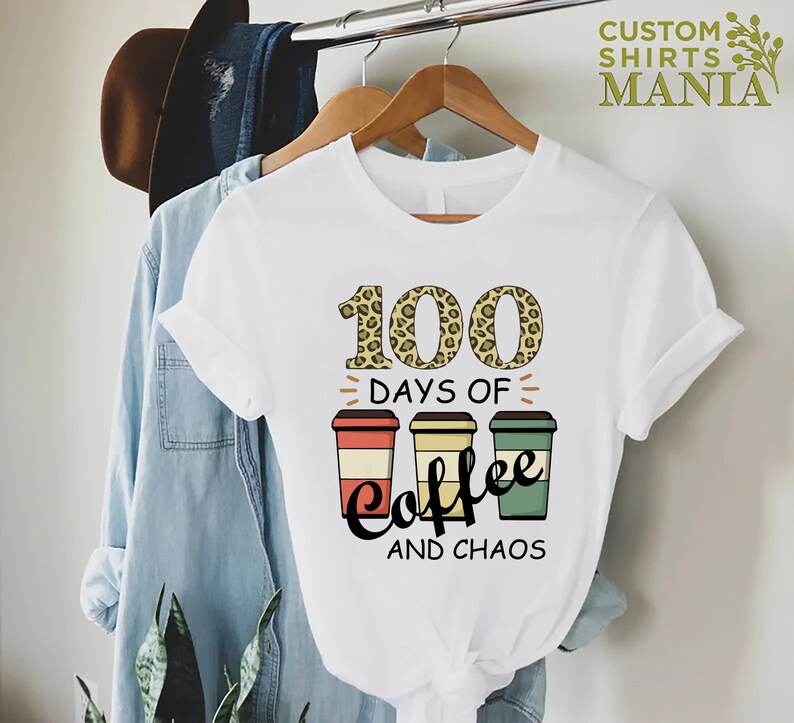 Pu&ograve; includere: Maglietta bianca con una stampa leopardata 100, il testo "Days of Coffee and Chaos" e tre tazze di caff&egrave; con strisce di diversi colori.