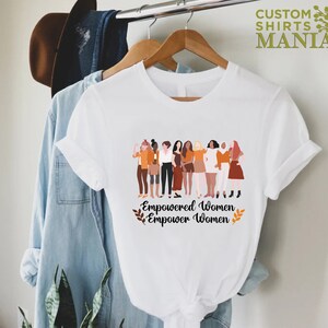 May include: White t-shirt featuring a graphic of a group of diverse women standing together. The text "Empowered Women Empower Women" is printed below the graphic.