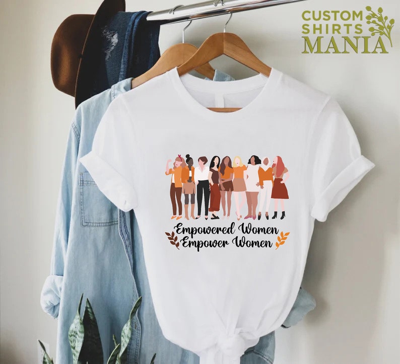 May include: White t-shirt featuring a graphic of a group of diverse women standing together. The text "Empowered Women Empower Women" is printed below the graphic.