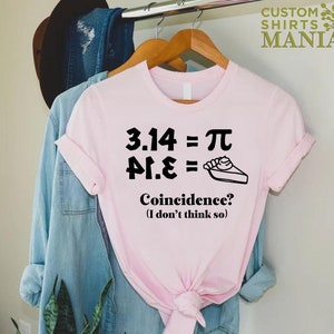 K&ouml;nnte beinhalten: Ein pinkfarbenes T-Shirt mit einer schwarzen Grafik, auf der "3.14 = &pi;, &pi;.14 =  " steht, sowie ein Bild von einem St&uuml;ck Kuchen. Der Text unter der Grafik lautet "Zufall? (Ich glaube nicht)"