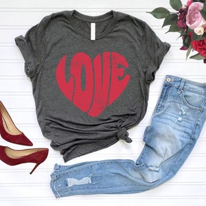 May include: A dark gray t-shirt with a red heart-shaped graphic that says "LOVE" in a distressed font. The shirt is paired with light blue jeans and red heels.